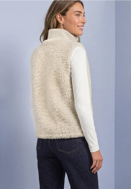 Femme en jean, col roulé blanc et gilet en moumoute beige, souriante et de profil légèrement.