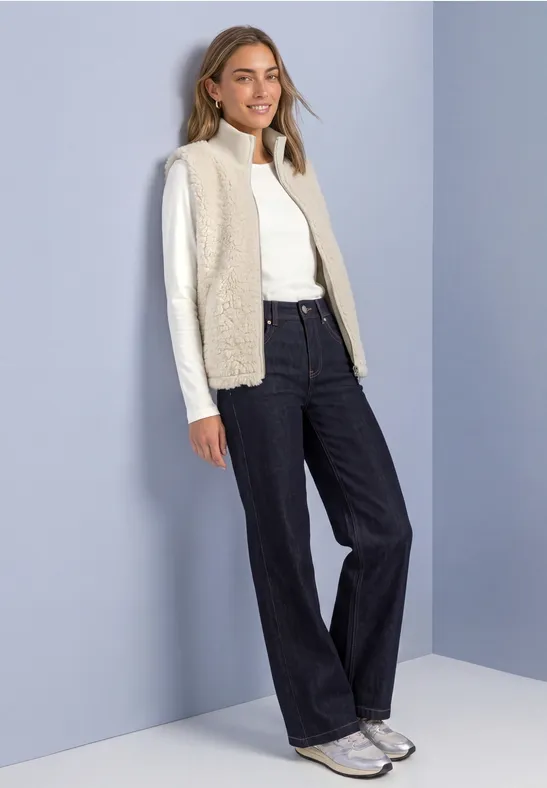 Femme en pull à col roulé blanc, gilet beige, jean large et baskets devant un mur pastel.