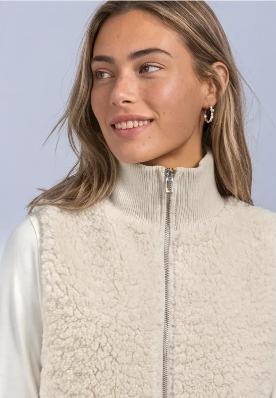 Jeune femme souriante portant une veste polaire beige et un haut à manches longues blanc.
