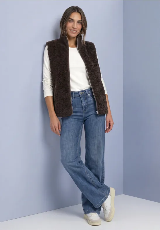 Femme porte chemise blanche, gilet en fausse fourrure brun foncé, jean large et baskets blanches.