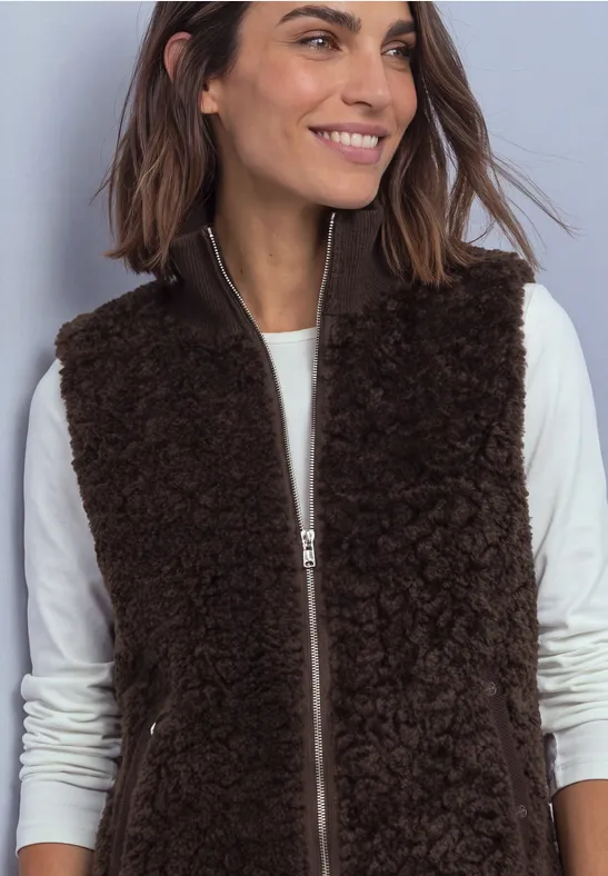 Femme en t-shirt à manches longues blanc porte une veste marron foncé et pelucheuse zippée, souriant.