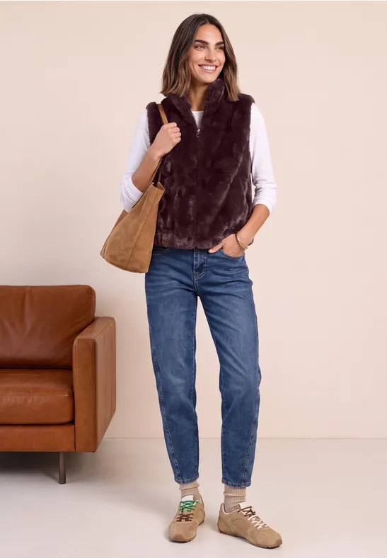 Jonge vrouw met witte blouse, bruine nep-bontjas, blauwe jeans, beige schoenen en bruine handtas.
