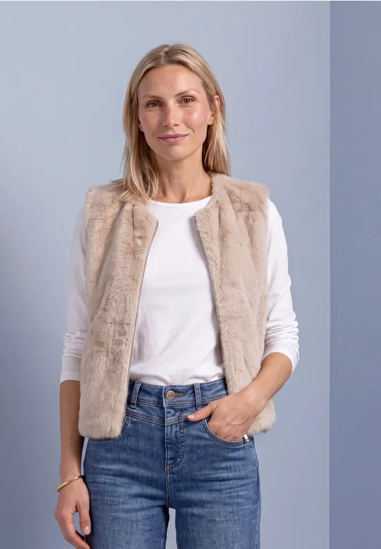 Eine Frau in weißem Shirt und beigem Fellweste, Jeans vor grauem Hintergrund.