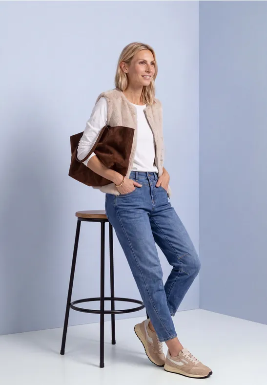 Eine lächelnde Frau in beigem Cardigan, weißem Shirt und Jeans lehnt an einem Hocker und trägt eine große Tasche.