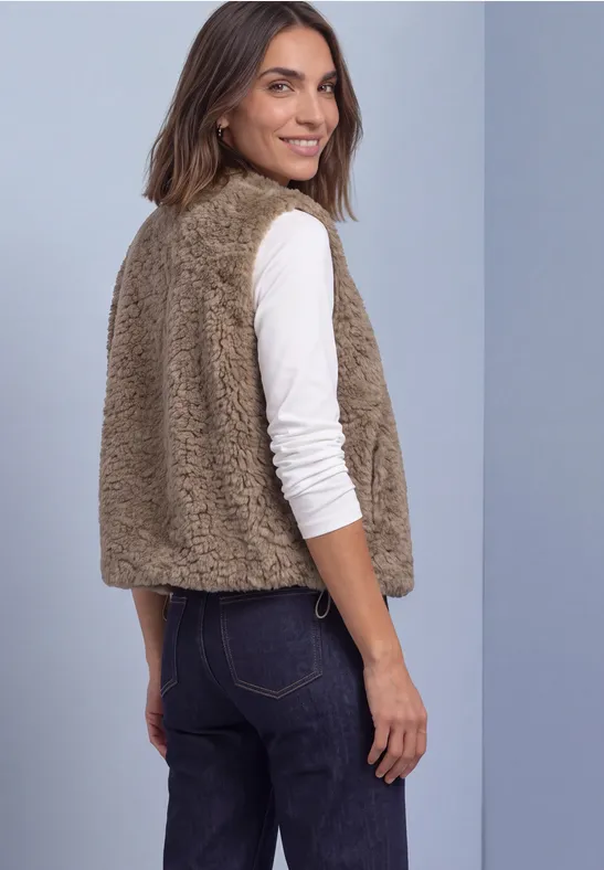 Eine Frau zeigt lächelnd von der Seite eine kamelfarbene Teddy-Weste über weißem Langarmshirt.