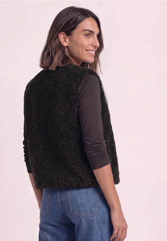 Vrouw in profiel draagt zwarte, pluizige bodywarmer over een longsleeve en jeans.