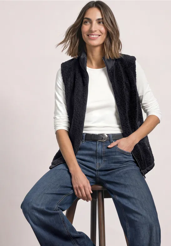 Vrouw zit op kruk, draagt zwarte teddy-vest, wit shirt en wijde jeans.