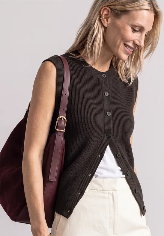Eine lächelnde Frau trägt eine schwarze ärmellose Weste, ein weißes Top, einen hellen Rock und eine bordeauxfarbene Tasche.