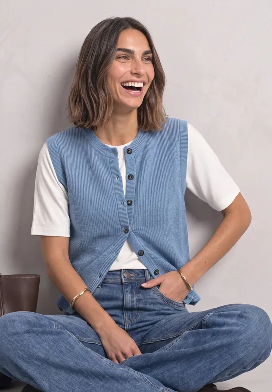 Eine lächelnde Frau sitzt im Schneidersitz, trägt eine weiße Bluse unter einer hellblauen Weste und blaue Jeans.