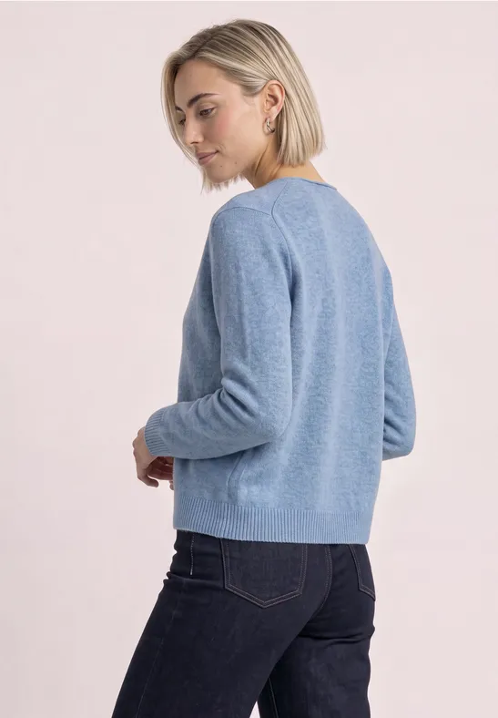 Eine Frau in blauer Strickjacke und dunklen Jeans, steht seitlich und schaut über die Schulter.