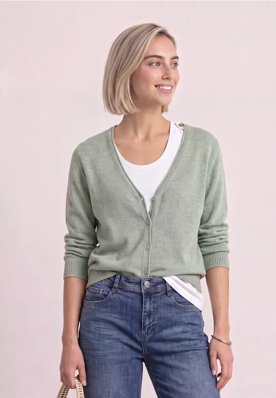 Junge Frau mit kurzem blondem Haar trägt hellgrüne Strickjacke, weißes Shirt und Jeans, sieht fröhlich aus.