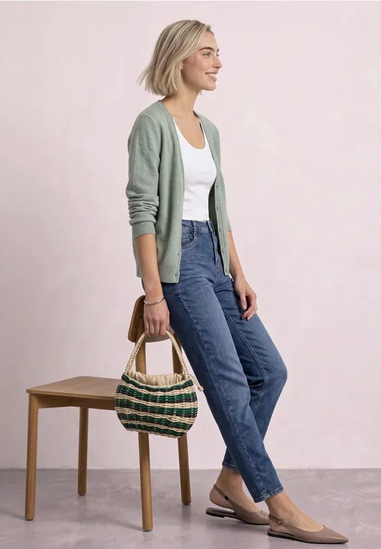 Frau mit grüner Strickjacke, weißem Top, blauen Jeans und Basttasche vor hellem Hintergrund.