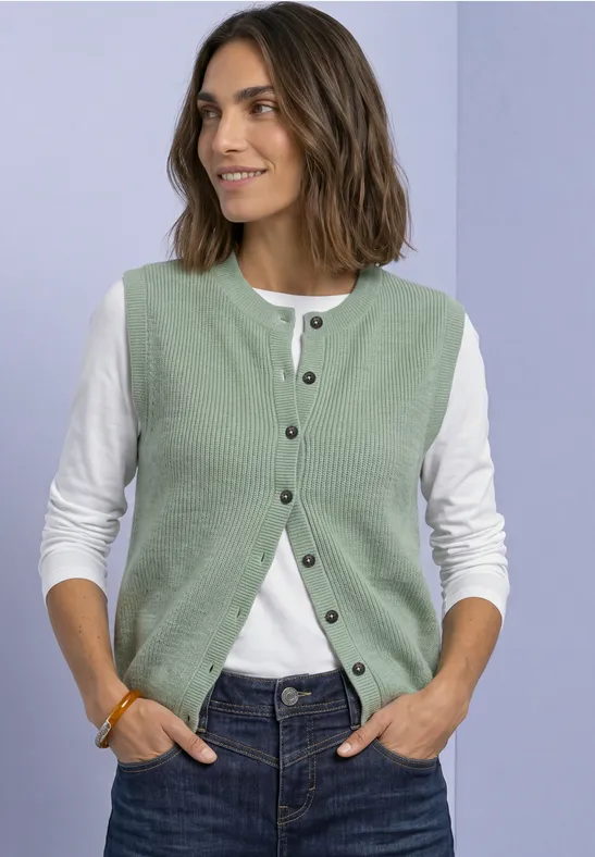 Frau in grüner Strickweste über weißem Langarmshirt und Jeans, lächelnd, Hände in den Taschen.