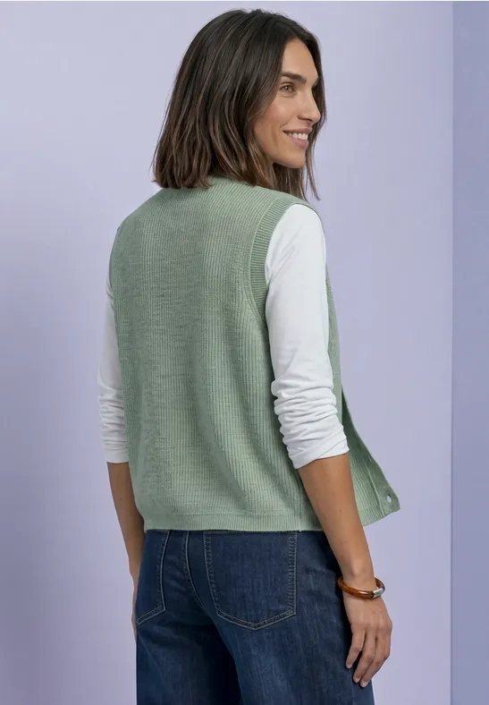 Eine Frau trägt eine grüne Strickweste über weißem Langarmshirt und Jeans vor lila Hintergrund.