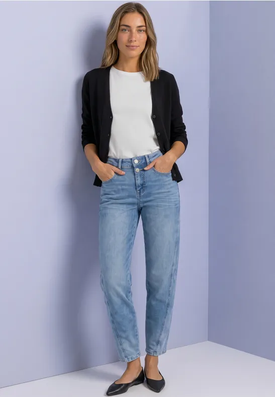 Eine lächelnde Frau im weißen Shirt, schwarzer Strickjacke und blauen Jeans vor lila Wand.