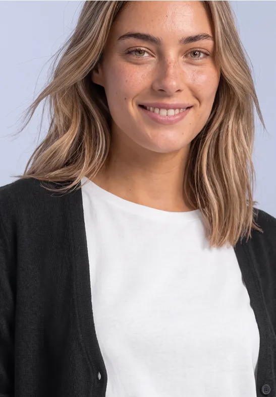 Eine junge Frau mit blonden Haaren und Sommersprossen lächelt in weißem T-Shirt und schwarzer Strickjacke.