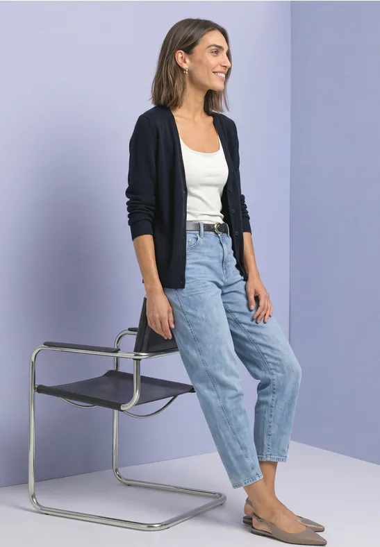 Femme en jean, débardeur blanc et cardigan, appuyée sur une chaise moderne devant un fond pastel.