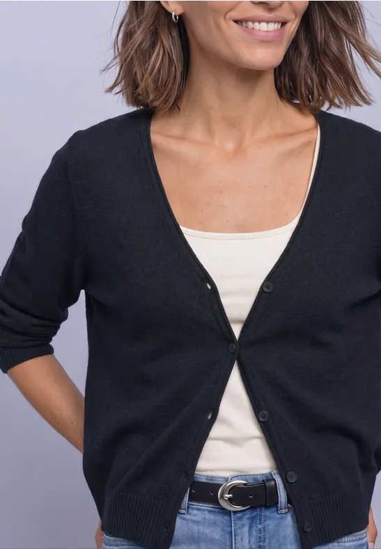 Buste d’une femme souriante en cardigan noir sur débardeur blanc, jean bleu et ceinture.