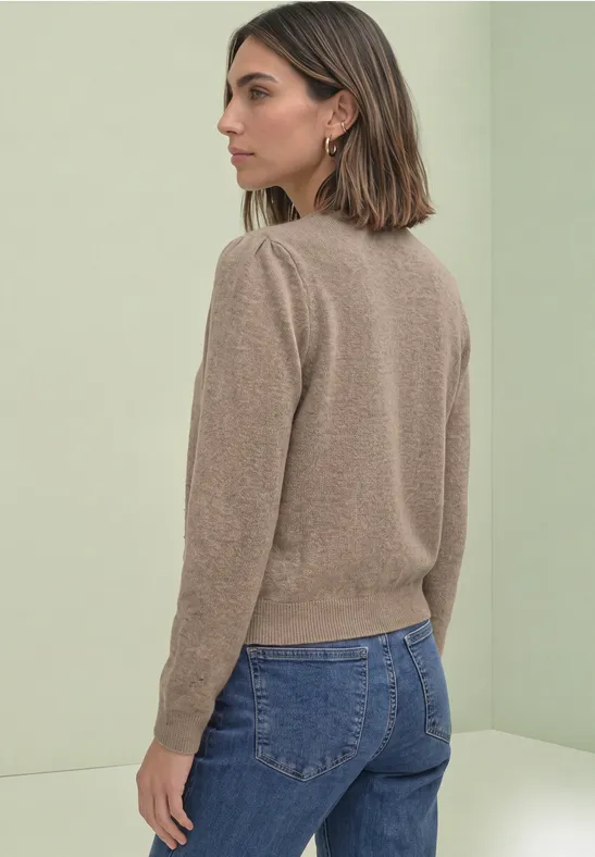 Vrouw met kort haar draagt een beige trui en blauwe jeans, zijwaarts poseerend voor lichtgroene achtergrond.