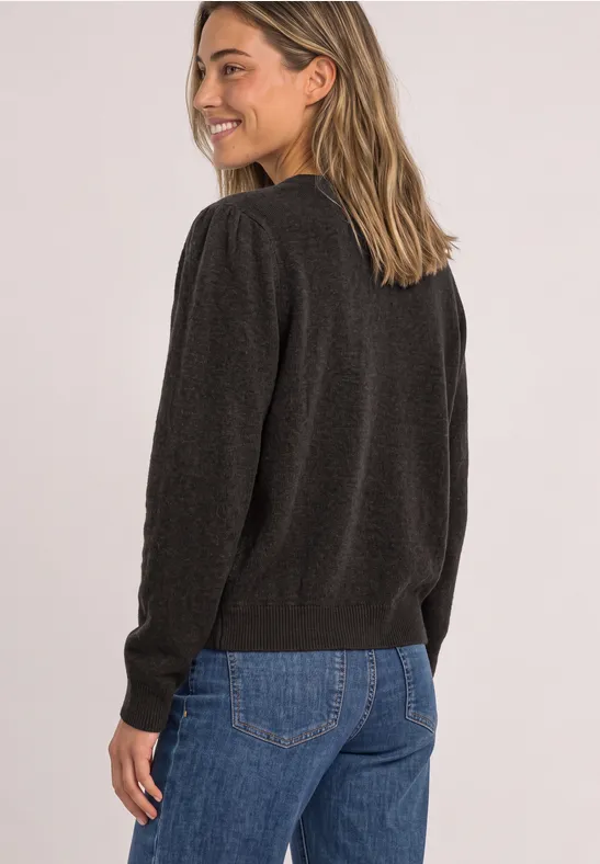 Eine Frau in schwarzem Pullover und blauer Jeans lächelt seitlich zur Kamera, mit Blick über die Schulter.