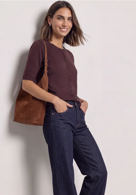 Eine Frau in braunem Cardigan und dunklen Jeans mit brauner Schultertasche lächelt vor neutralem Hintergrund.