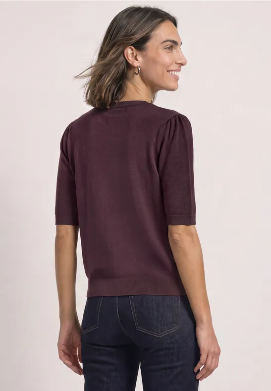 Frau mit bordeauxfarbigem Kurzarmpullover und dunkler Jeans, Rücken zur Kamera, seitlich schauend.