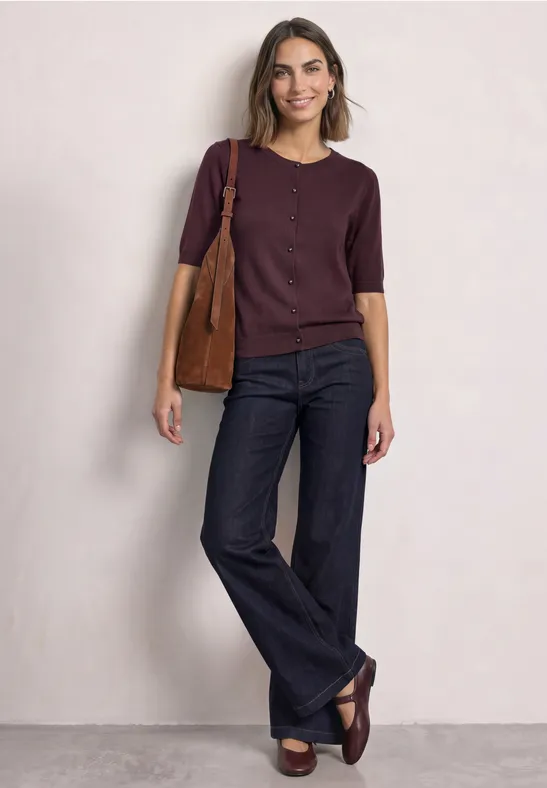 Frau in lila Oberteil und ausgestellten Jeans, trägt Tasche und locker lächelnd.