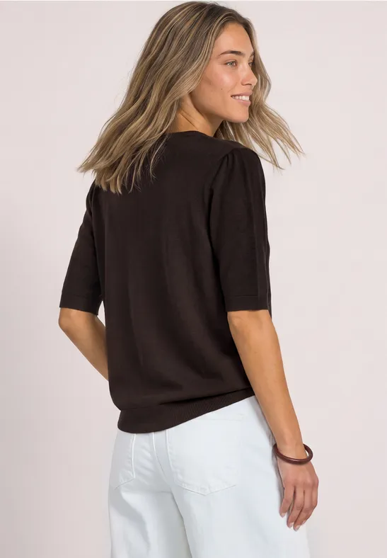 Junge Frau im schwarzen Kurzarm-Pullover und weißen Hosen, seitlich von hinten, lächelnd vor hellem Hintergrund.