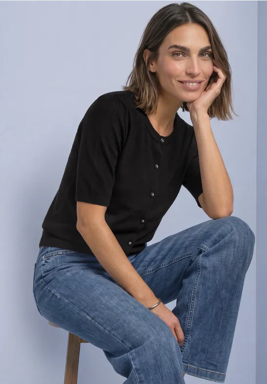 Eine lächelnde Frau in schwarzem Shirt und blauen Jeans sitzt entspannt auf einem Hocker.