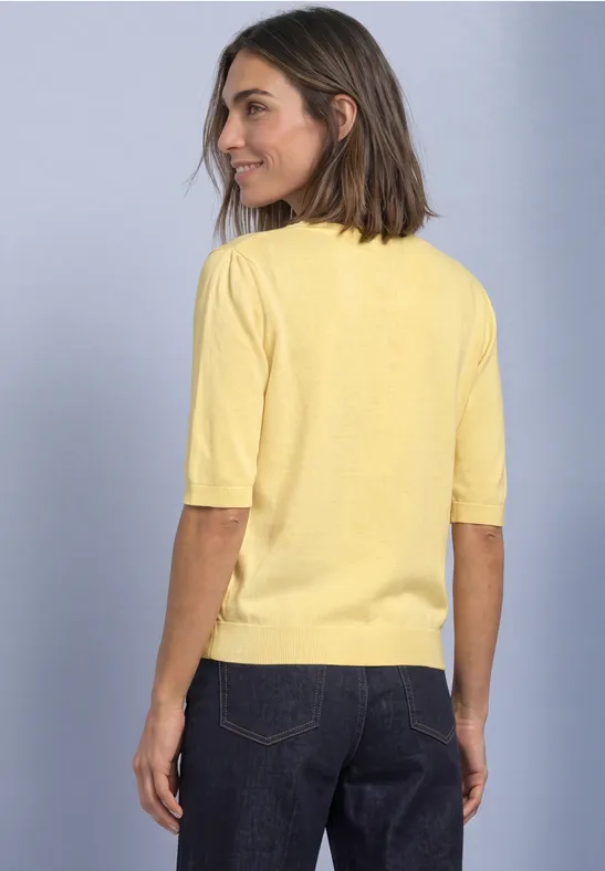 Femme de dos vêtue d’un haut jaune à manches courtes et d’un jean foncé sur fond neutre.