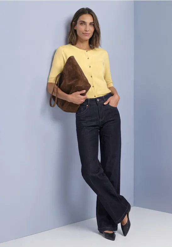 Femme en chemise jaune et jean sombre, tient un sac marron et s’appuie décontractée contre un mur.
