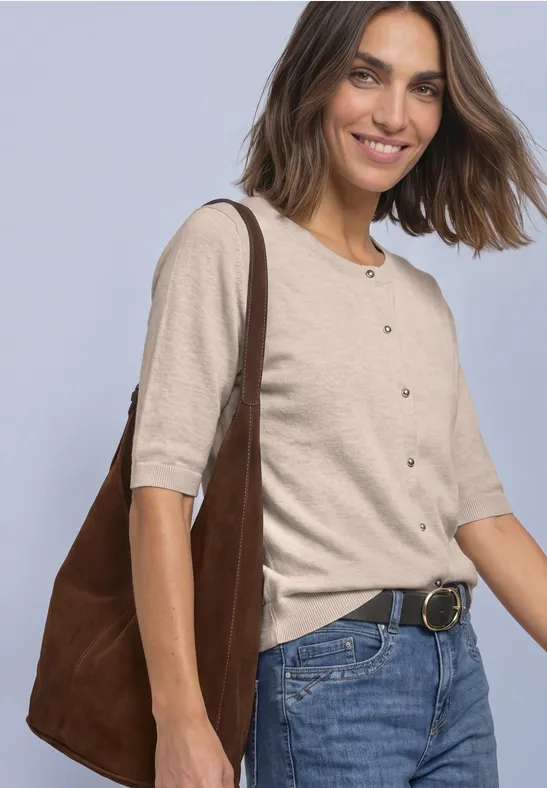 Lächelnde Frau in Jeans, beiger Kurzarm-Strickjacke und brauner Schultertasche vor hellem Hintergrund.