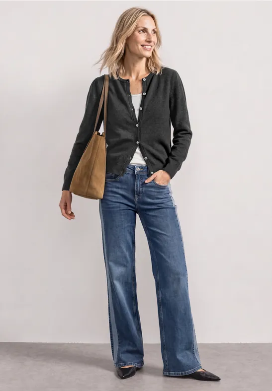 Eine Frau trägt schwarze Strickjacke, weite Jeans, flache Schuhe und eine braune Tasche.