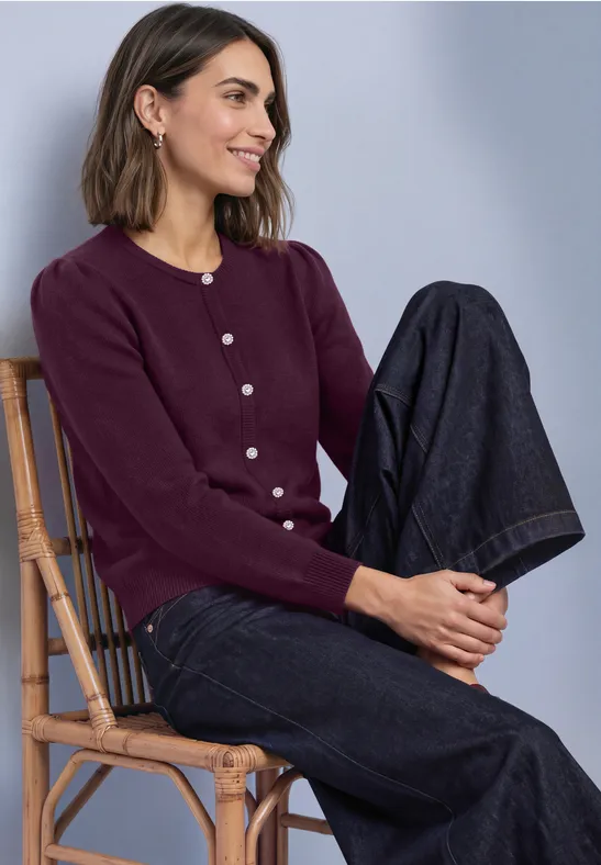 Eine lächelnde Frau sitzt auf einem Korbstuhl, trägt einen lila Cardigan mit Knöpfen und weite Jeans.