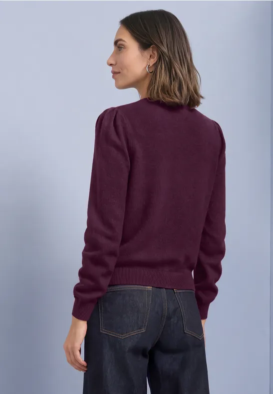 Eine Frau in Profilansicht trägt einen dunkelroten Pullover und dunkle Jeans vor neutralem Hintergrund.