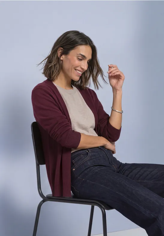 Eine lächelnde Frau sitzt auf einem Stuhl, trägt einen weinroten Cardigan, beige Top und blaue Jeans.