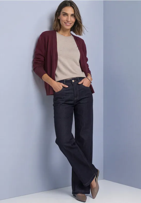 Frau lehnt lässig vor pastellfarbenem Hintergrund, trägt weinrote Strickjacke, beiges Top, dunkle Jeans und Pumps.