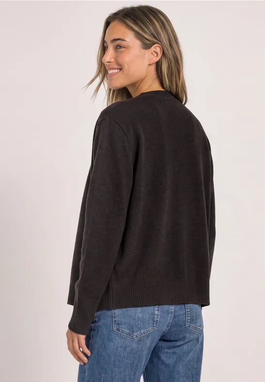 Frau im schwarzen Cardigan und blauen Jeans, von hinten fotografiert und über die Schulter blickend.