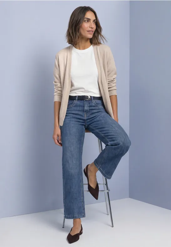 Femme portant un t-shirt blanc, un cardigan beige, un jean droit bleu et des ballerines brunes devant un mur clair.