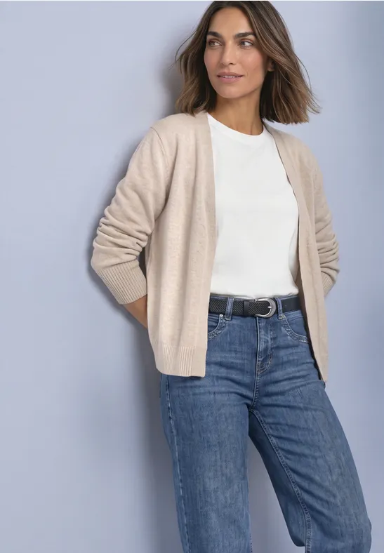 Femme décontractée portant un gilet beige, un t-shirt blanc et un jean devant un mur clair.