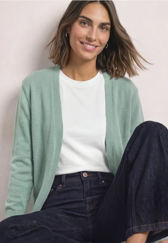 Frau sitzt lächelnd, trägt grüne Strickjacke, weißes T-Shirt und dunkle, weite Jeans.