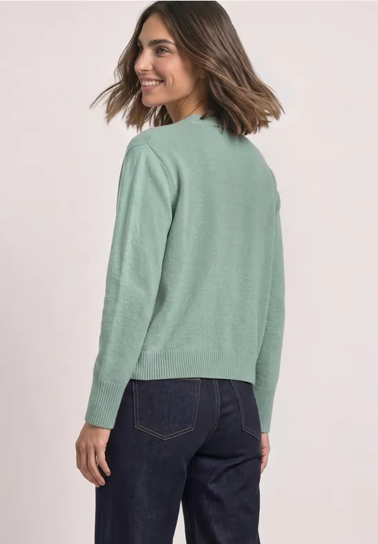 Eine Frau trägt einen hellgrünen Pullover und dunkle Jeans, blickt lächelnd über die Schulter.