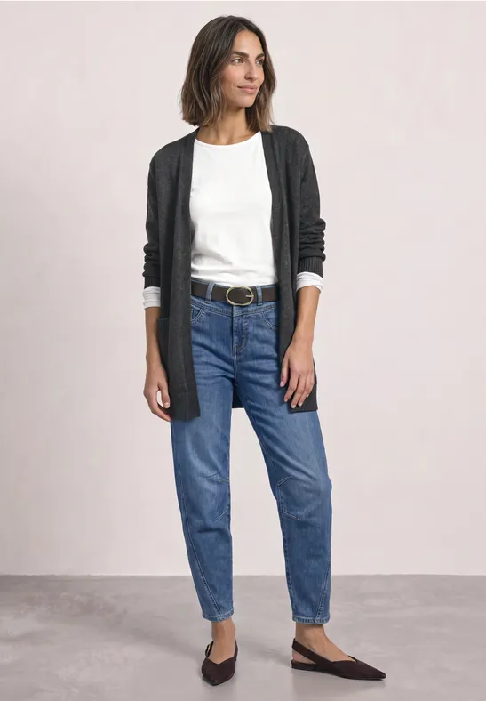 Eine Frau trägt schwarze Strickjacke, weiße Bluse, blaue Jeans und schwarze Ballerinas vor neutralem Hintergrund.