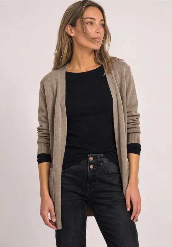 Eine Frau in einem langen beigen Cardigan, schwarzem Oberteil und dunkler Jeans steht vor hellem Hintergrund.