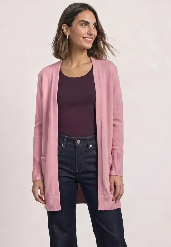 Frau trägt rosa Strickjacke über dunklem Top und Jeans, lächelt selbstbewusst.