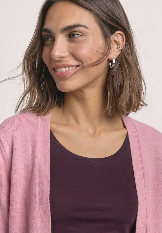 Eine lächelnde Frau mit braunen Haaren trägt ein dunkles Top und einen rosafarbenen Cardigan.