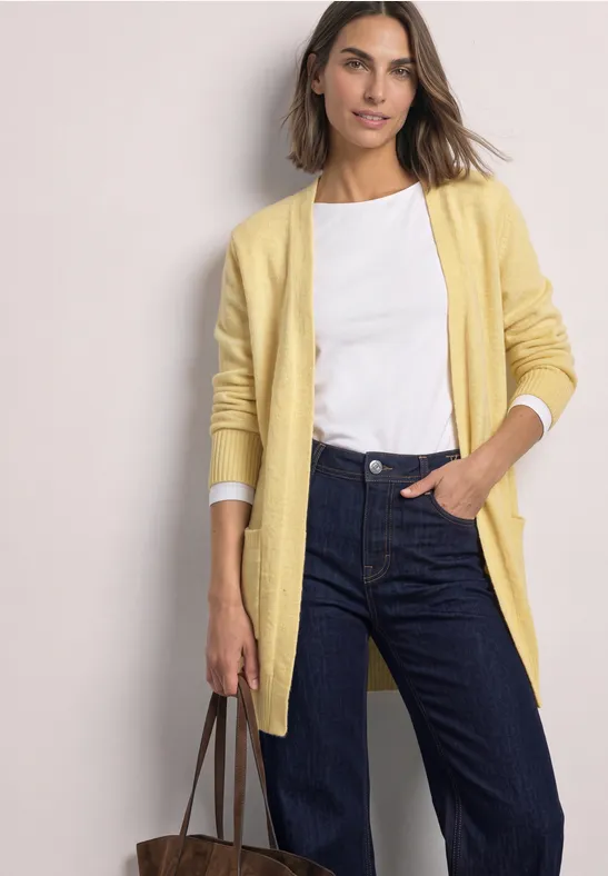 Eine Frau trägt weiße Bluse, gelben Cardigan und blaue Jeans, hält Ledertasche.