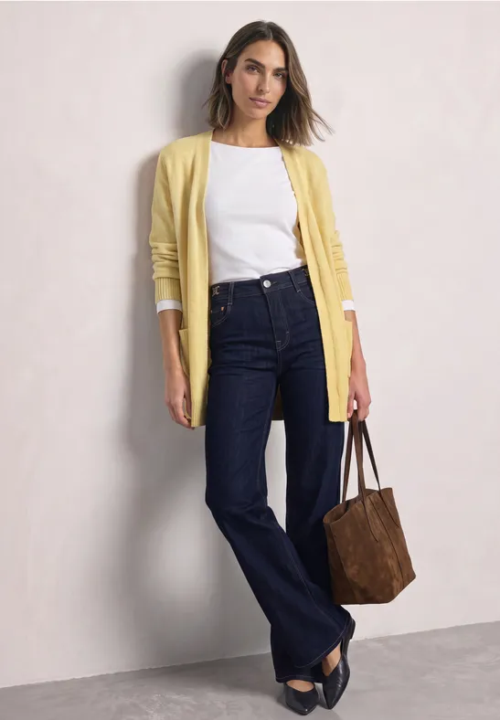 Frau in weißem Top, gelbem Cardigan, dunklen Jeans und brauner Tasche vor hellem Hintergrund.