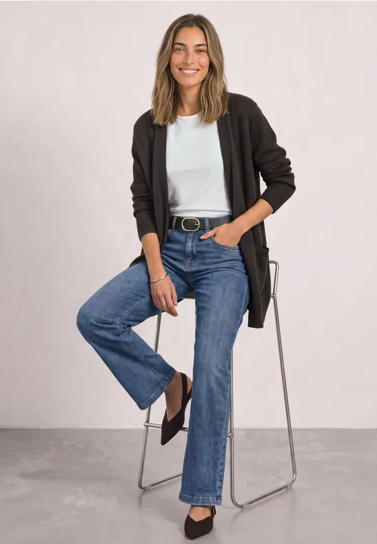 Femme assise sur un tabouret, portant un t-shirt blanc, un cardigan noir et un jean bleu, souriante.