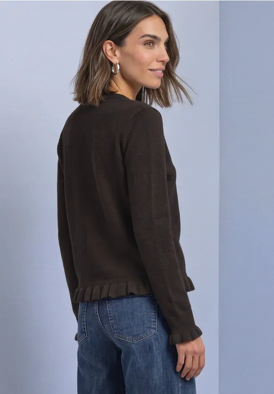 Frau seitlich stehend in dunkelbraunem Pullover mit Rüschenabschluss und blauer Jeans vor hellgrauem Hintergrund.